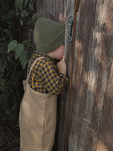 child in green beanie gingham flannel shirt & terry cotton beige overalls peeking through wooden door