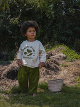 child in pea pod green wide pants & cream sweatshirt with logo standing in garden beside straw basket & fruits