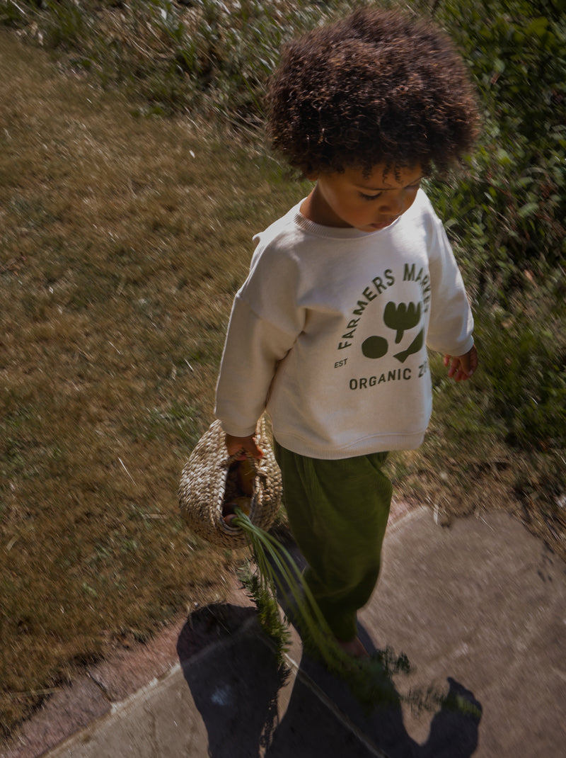 child dressed in cream sweatshirt with logo & green wide leg pants carrying straw basket with carrots