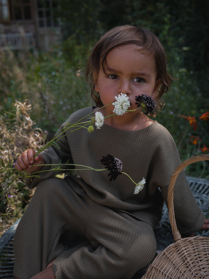 child dressed in matching cotton waffle olive sweatshirt & pants sitting on patio table smelling flowers