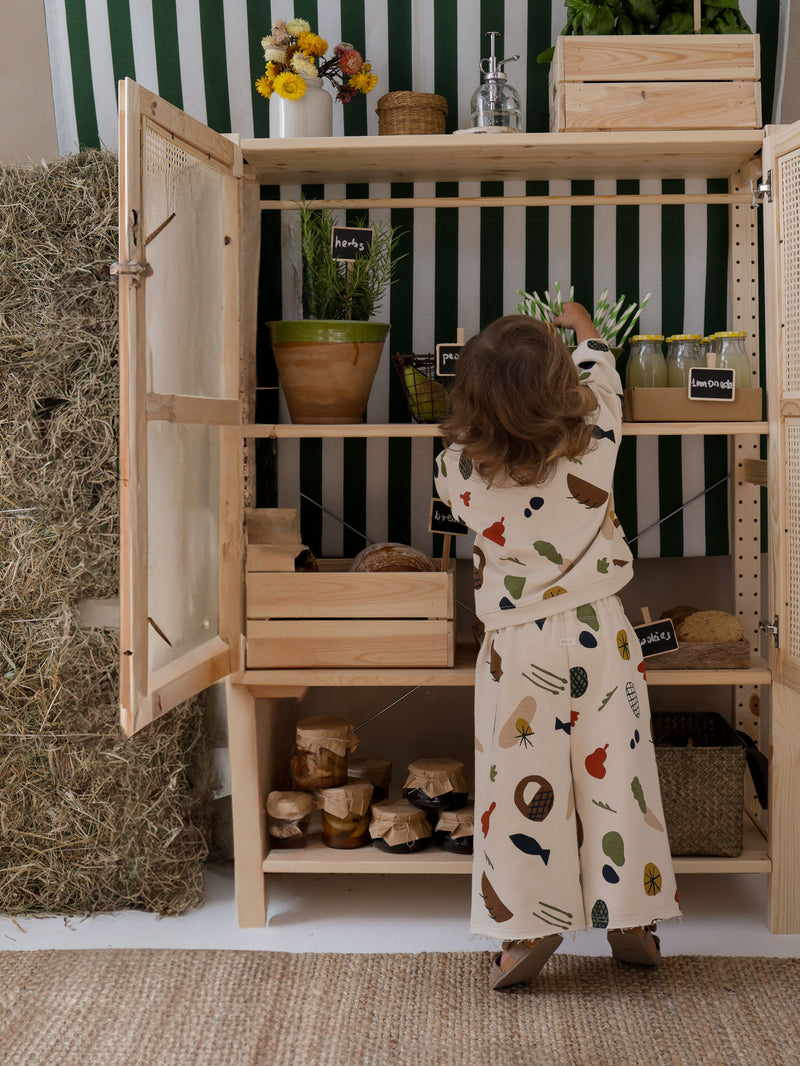 child in wide long pants & sweatshirt with farm produce print in tiptoes reaching for straw in cupboard