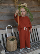 child with big scrunchie & red boxy sweatshirt pants set with white drawstring holding up a carrot