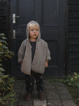 child in oatmeal wool jacket with hood over head & black sweatshirt & sweatpants standing in front of door