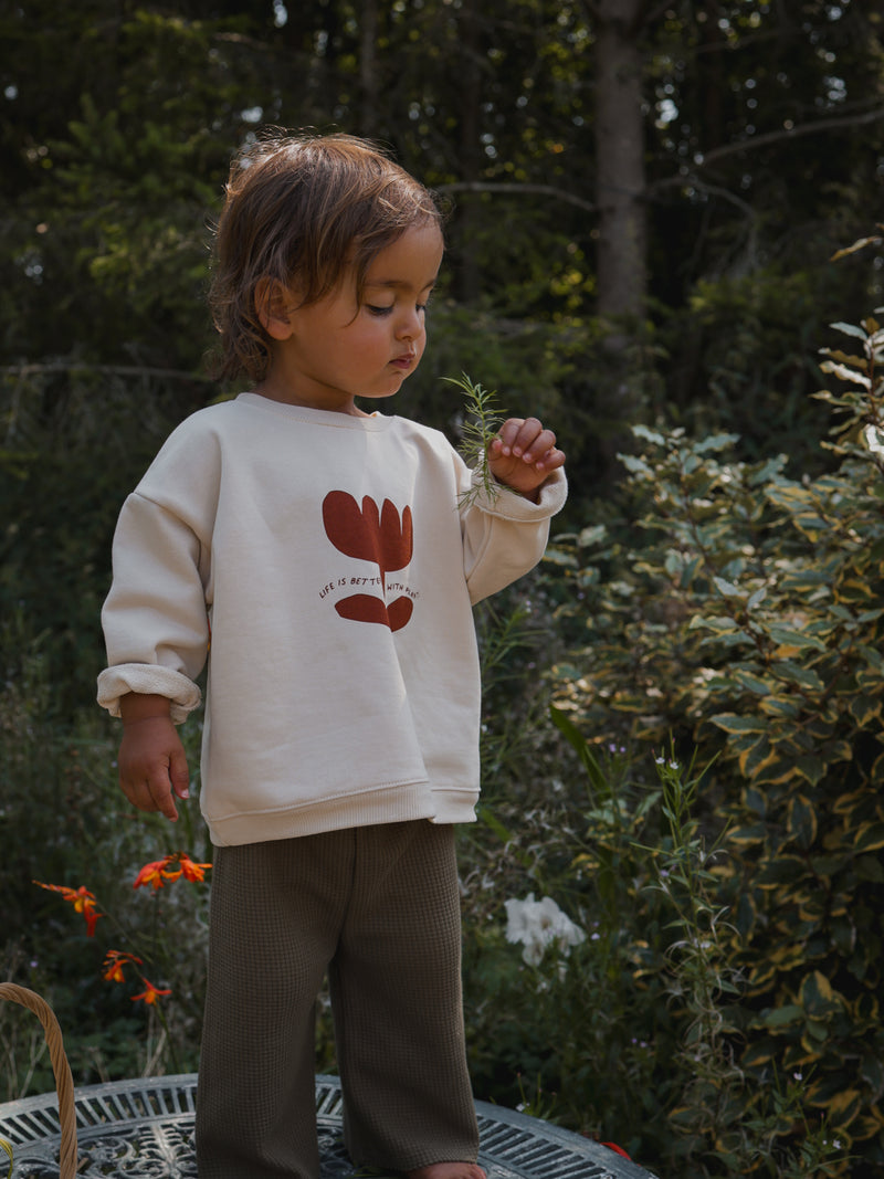 child in oversized cream sweatshirt & wide long pants holding herbs standing on patio table