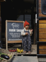 child in red beanie cream sweatshirt & oversized dungarees standing in front of farm shop a-frame sign