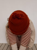 child in red beanie with tassel cream sweatshirt & red stripes leggings with head between knees