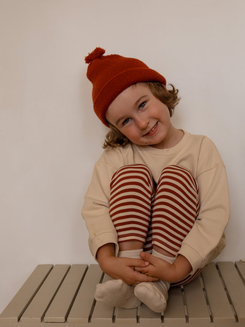 child wearing ribbed cotton leggings with red stripes cream sweatshirt & beanie sitting down with knees to chest