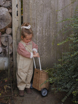 child with bun hairdo wearing red stripes sweatshirt with grey overalls pulling rattan trolley