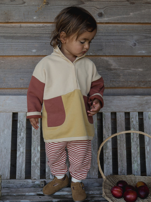 child in paprika red stripes long pants & colour block sweater standing beside rattan basket with apples