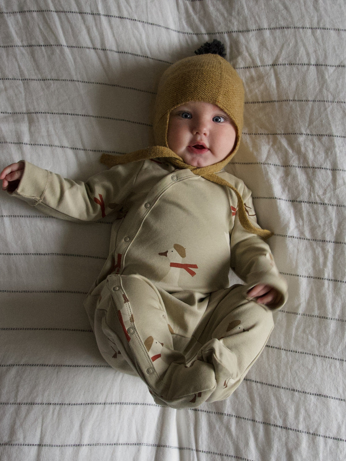 A baby lying on a striped white blanket, wearing a beige footed onesie with small red and brown animal prints and a mustard yellow knit bonnet with a pom-pom.