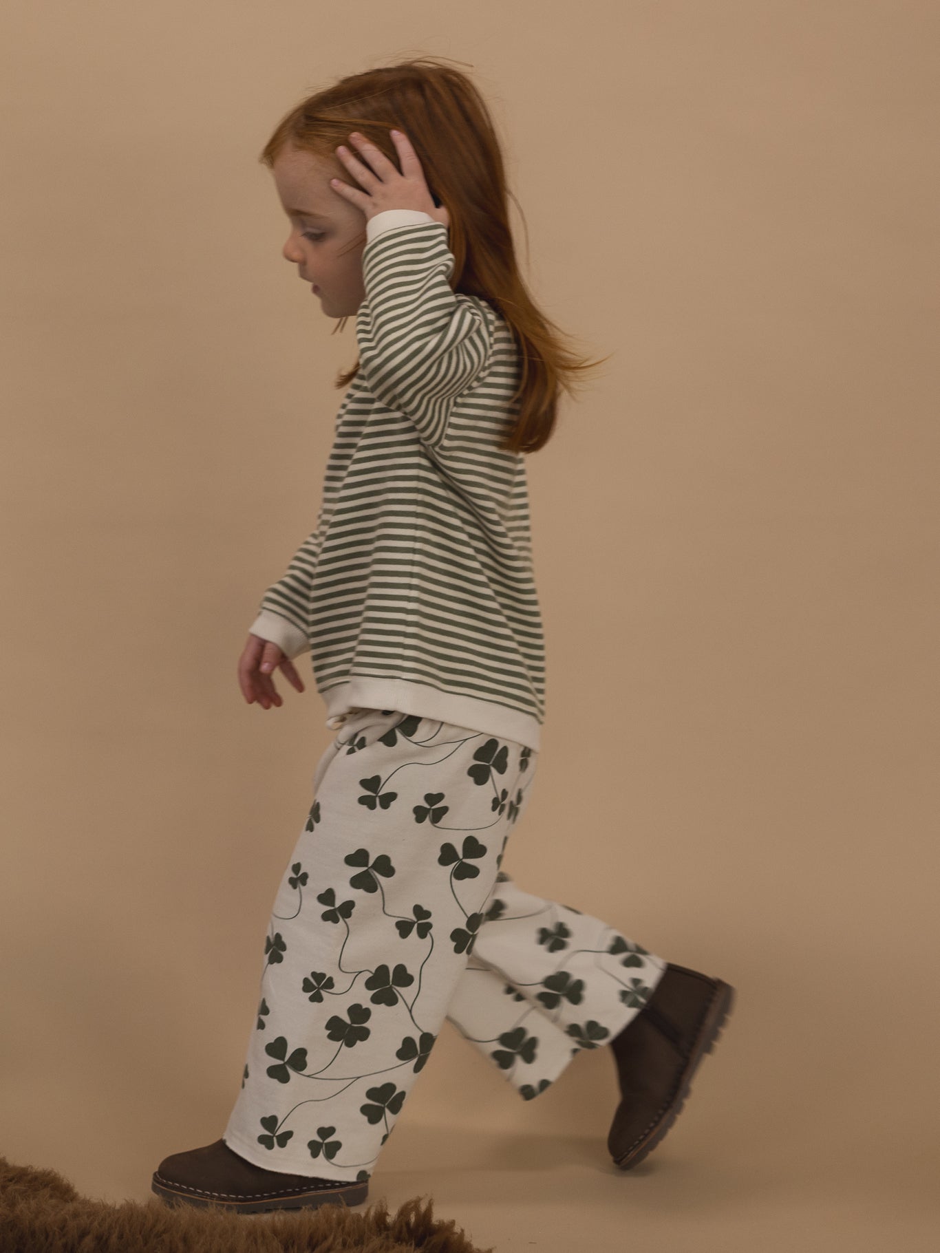 toddler in wide leg pants with green clover print and green striped sweatshirt, brushing hair away from face