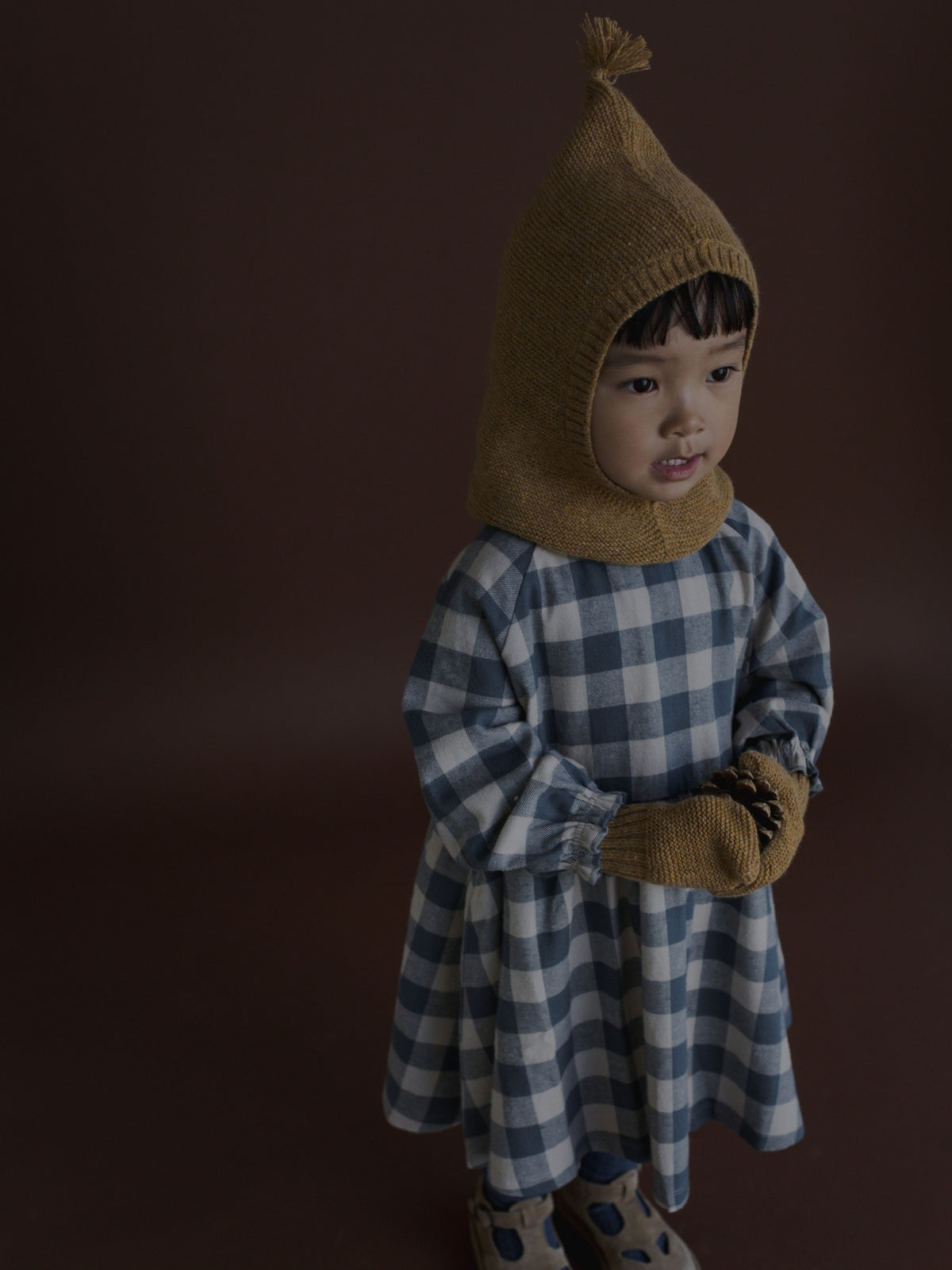 A toddler standing against a dark brown studio background, wearing a brown knit hooded hat with a tassel, a blue and white gingham dress, and matching brown mittens.