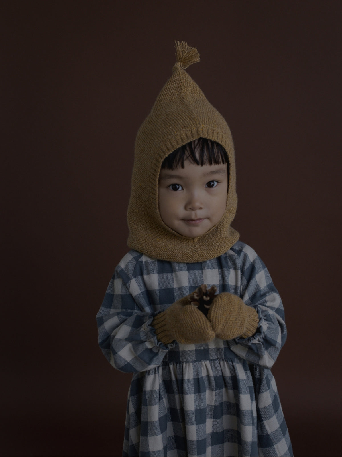 A toddler wearing a brown knit hooded outfit with a blue and white checkered dress, standing against a dark brown studio background.