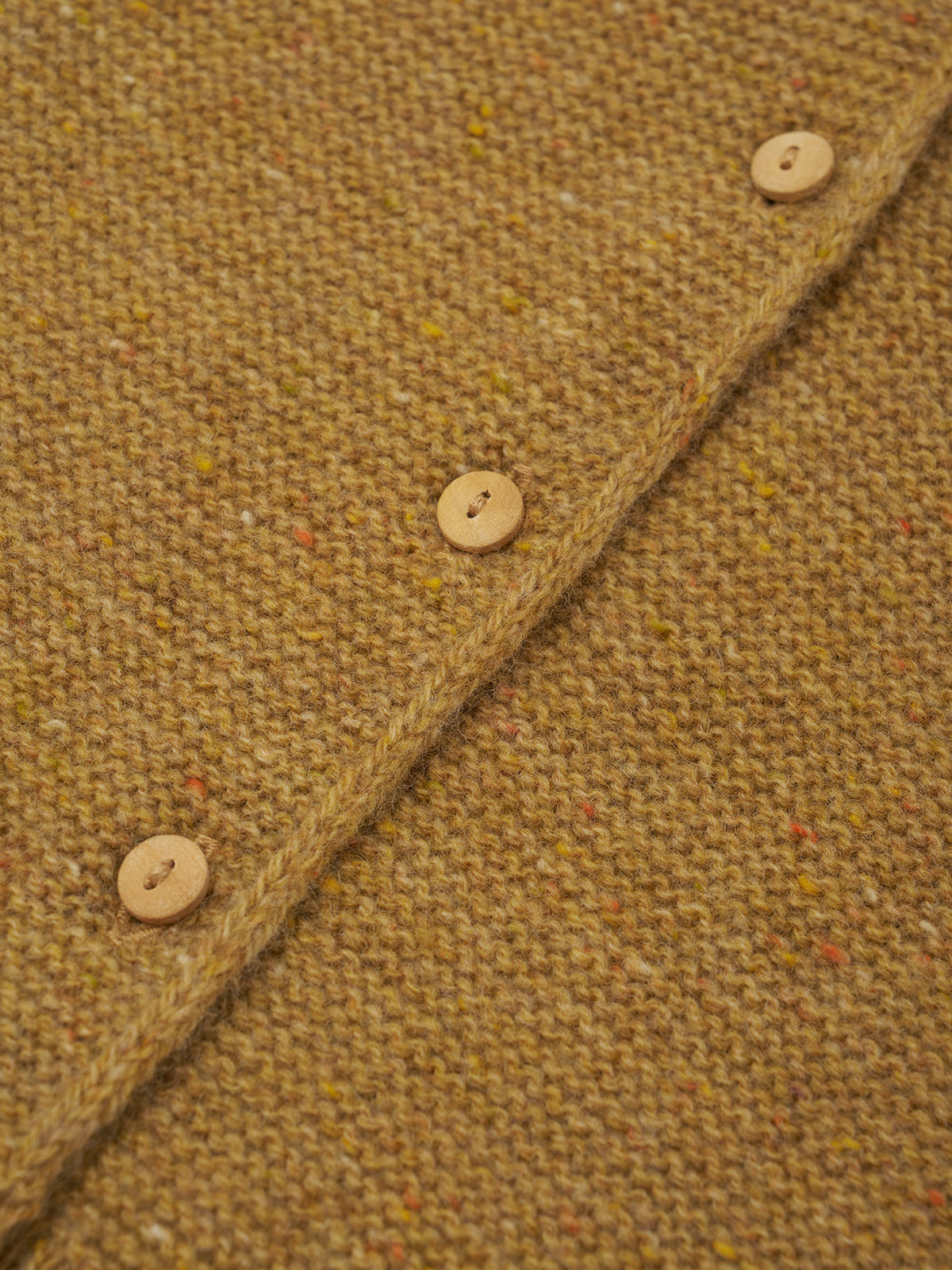 Close-up of a textured woven fabric in a warm camel-brown tone with small multicolored flecks, featuring a diagonal buttoned seam and light wood buttons.