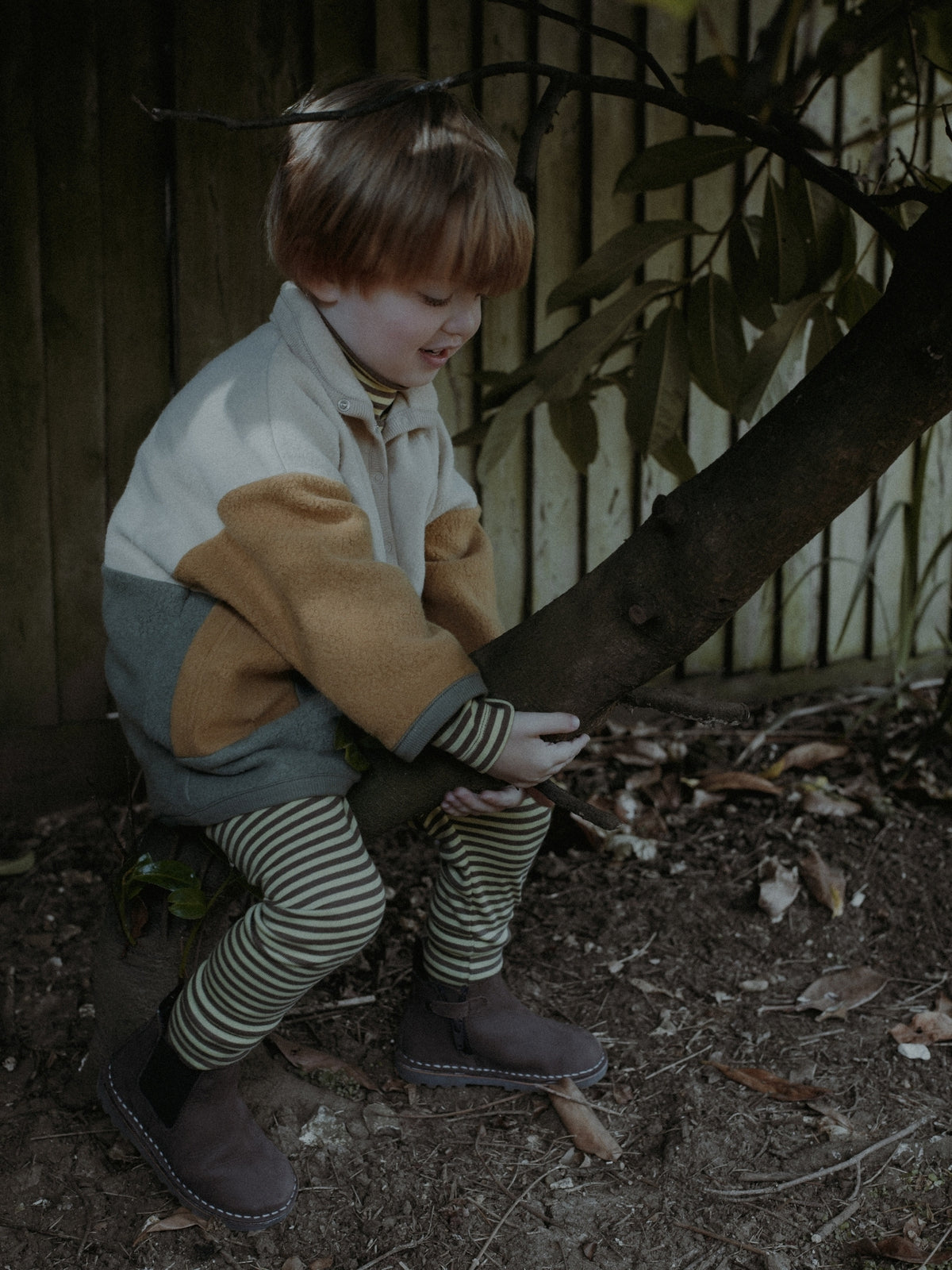 A young child wearing a color-blocked jacket with beige, brown, and gray panels, striped tights, and brown boots, sitting on the ground next to a tree trunk in an outdoor backyard setting.