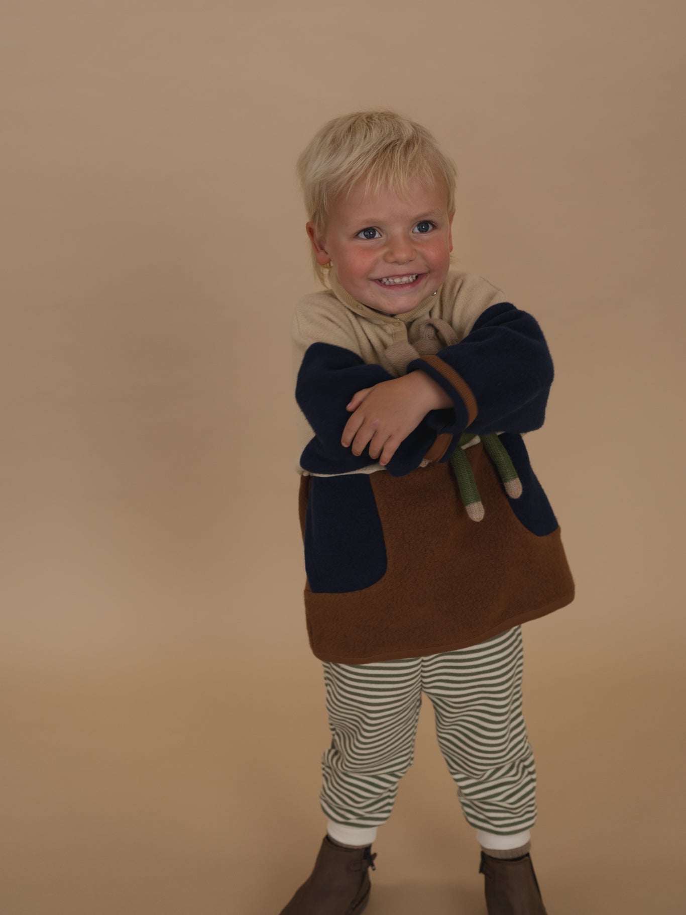 toddler wearing cream, navy and brown colour block fleece sweater with striped pants, hugging a plushie
