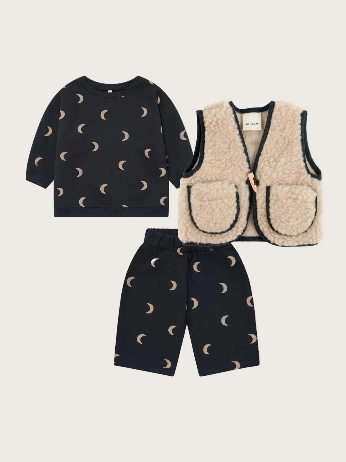 A three-piece baby clothing set featuring a black long-sleeve top and matching shorts with a beige crescent moon pattern, paired with a beige sherpa vest with black trim.