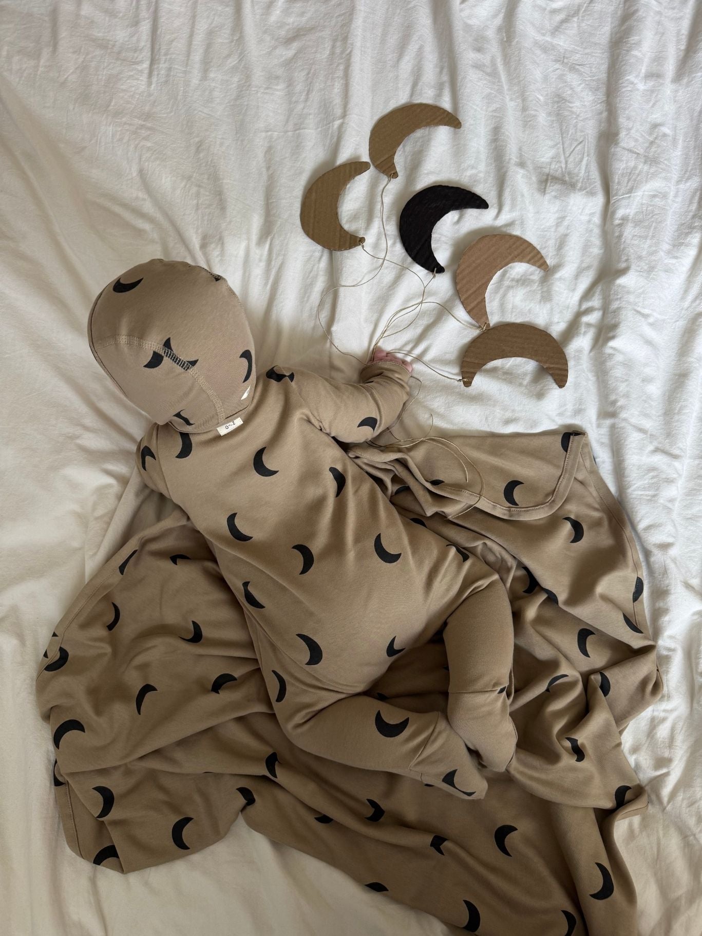 A baby wearing a beige hooded one-piece outfit with a black crescent moon print, lying on white bedding with matching moon-patterned blankets and small crescent moon toys nearby.
