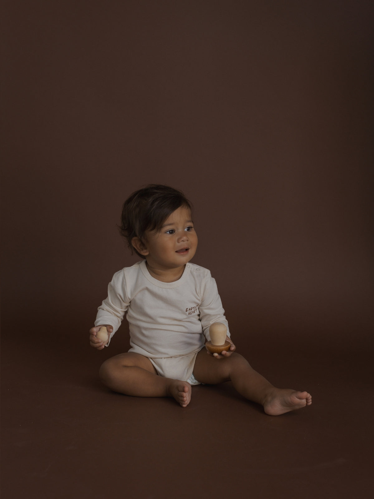 A baby sitting on a dark brown studio background, wearing a white long-sleeve bodysuit and holding a small wooden toy.