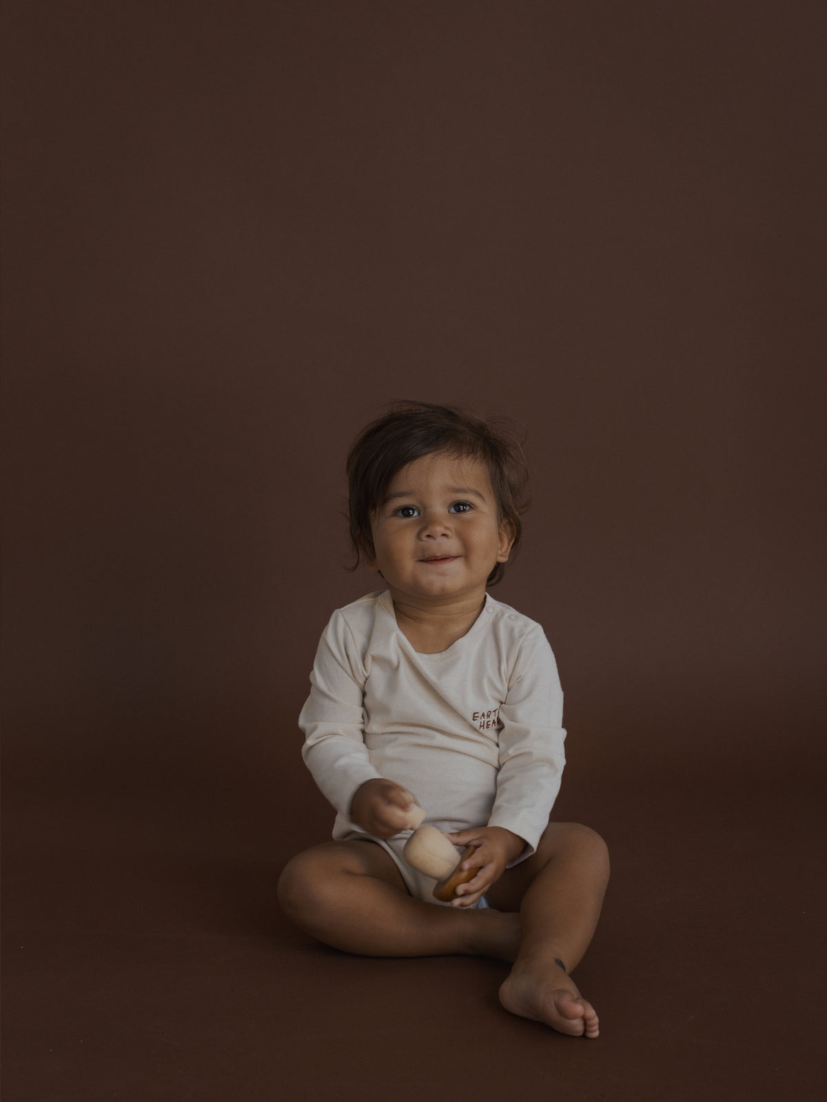 A baby sitting cross-legged on a dark brown studio background, wearing a cream long-sleeve bodysuit and holding a small beige toy.