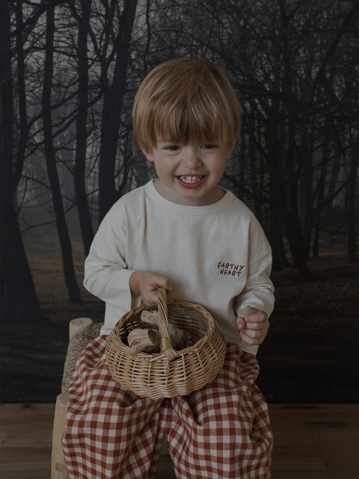 A smiling young child sits on a wooden stool holding a small wicker basket, wearing a cream long-sleeve shirt with 'EARTHY HEART' text and red-and-white gingham pants, against a dark forest backdrop.