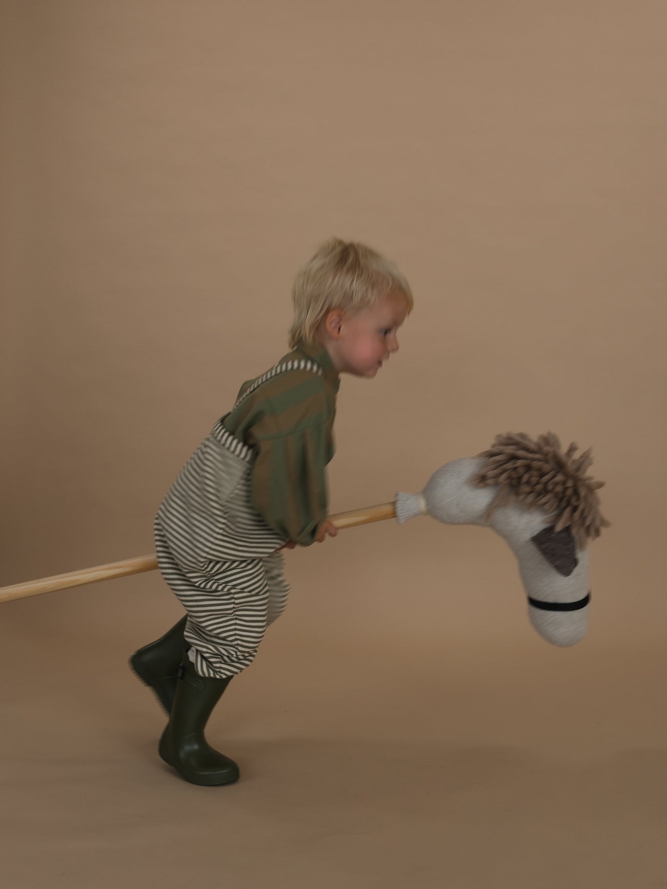 toddler in green and white striped dungarees with lodge shirt and rain boots, galloping on hobby horse