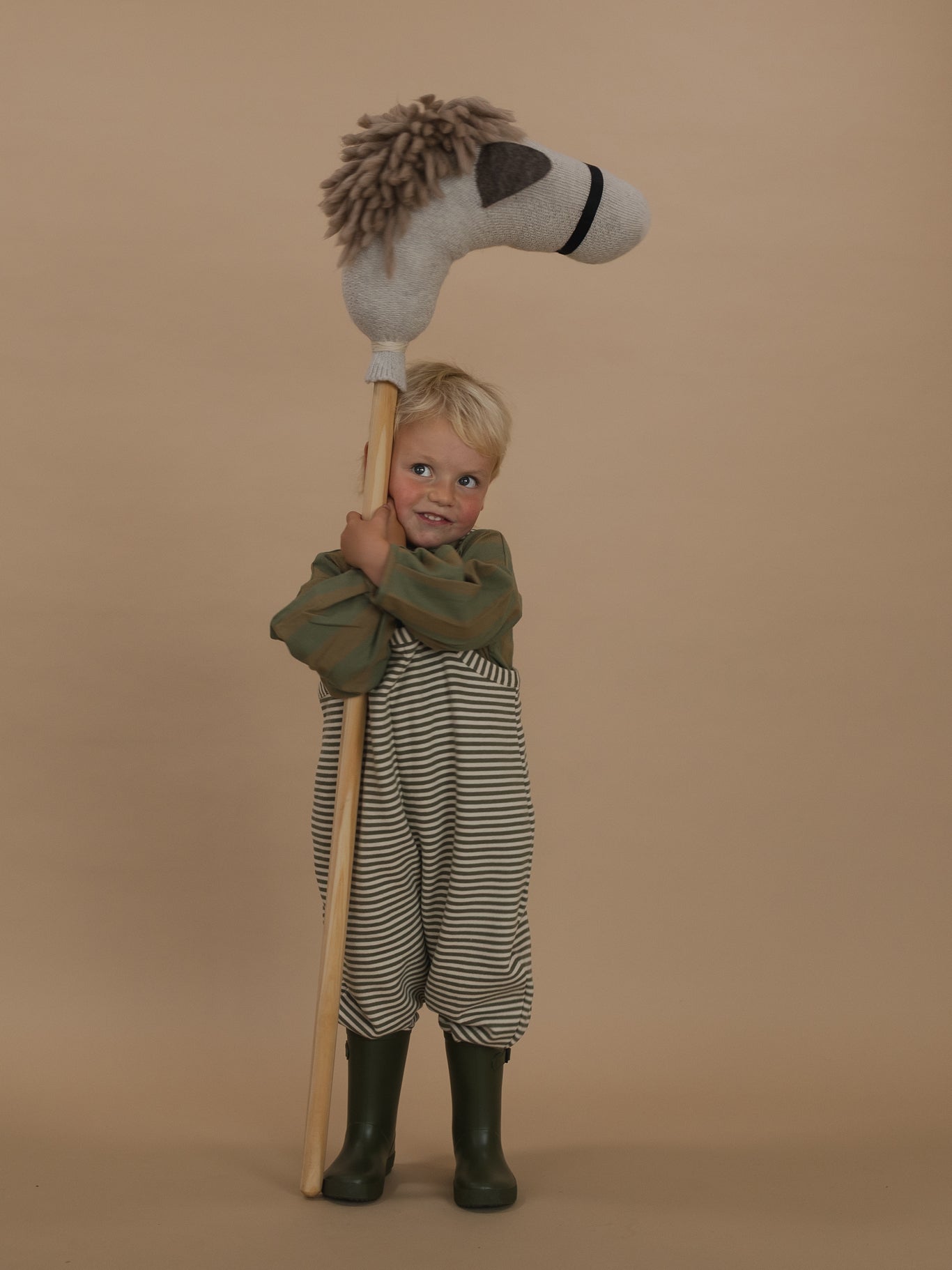 toddler in green and white striped dungarees with lodge shirt and rain boots, hugging a hobby horse