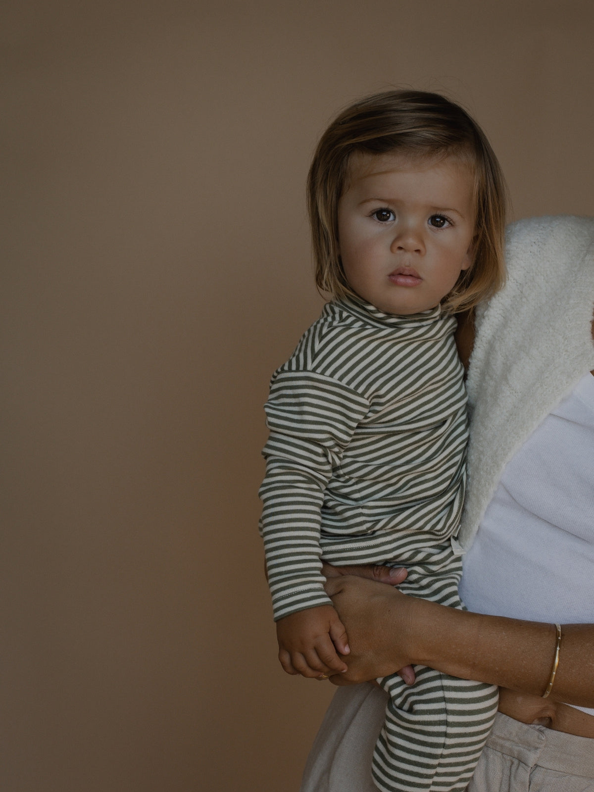 A toddler wearing a muted green and beige striped long-sleeve turtleneck jumpsuit, held by an adult against a solid warm brown studio background.