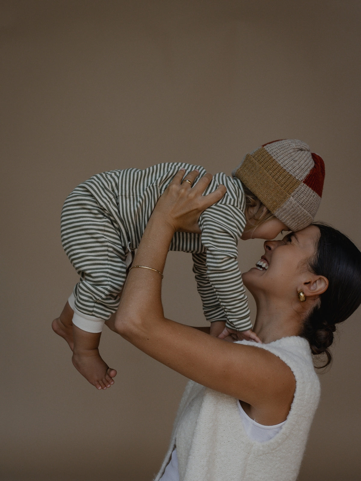 A woman holds a baby up in the air against a plain beige studio background. The baby is wearing a green and white striped one-piece outfit and a beige and red knit beanie.