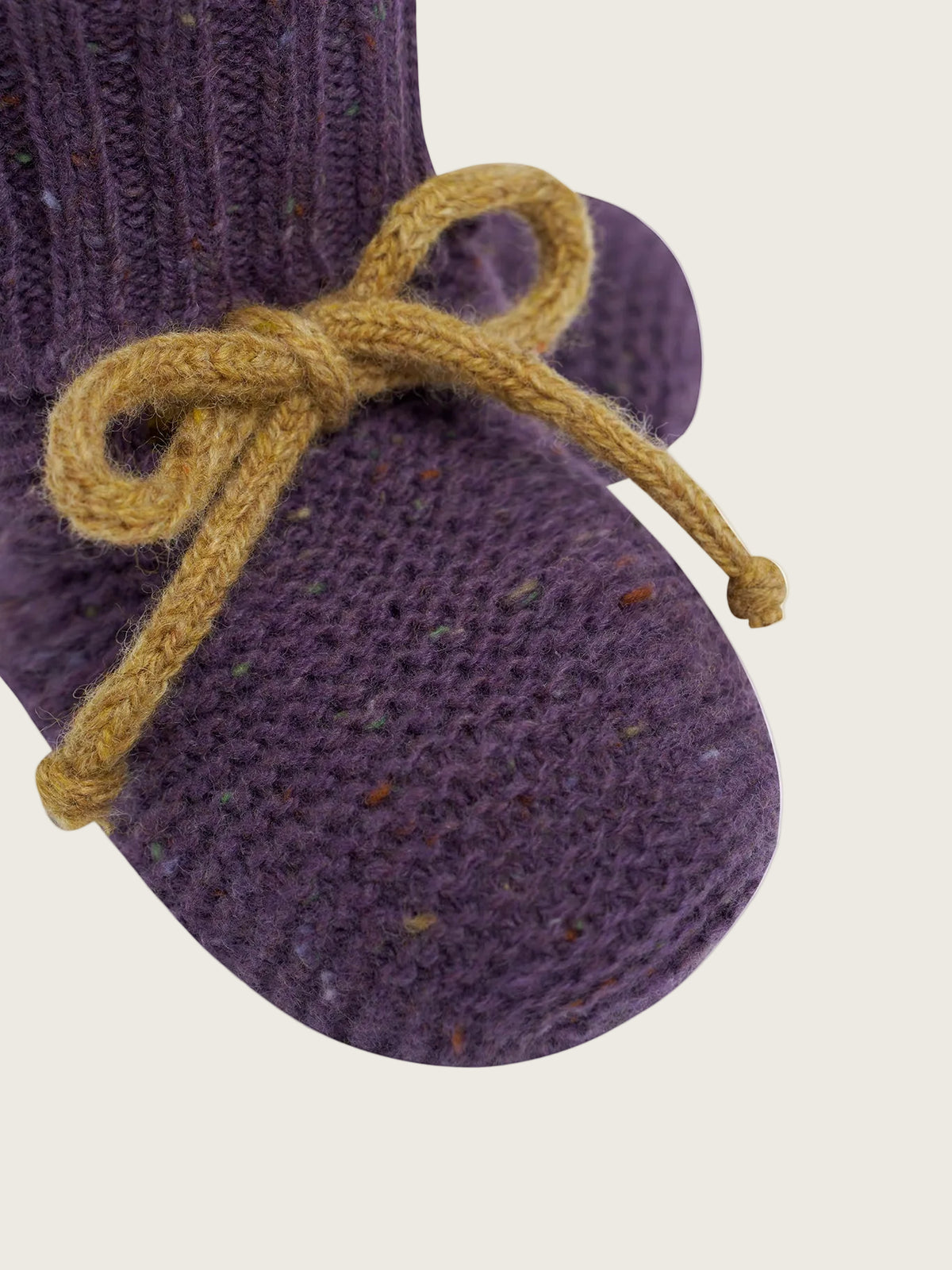 A close-up of a purple knit bootie with a mustard yellow knotted bow, set against a solid off-white background.