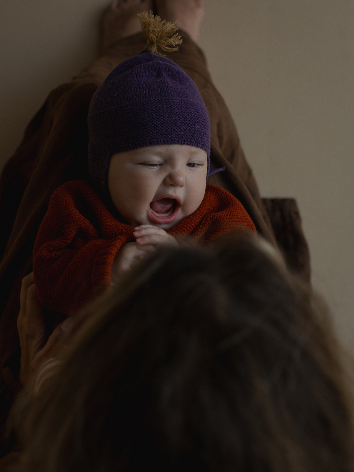 A baby wearing a purple knit hat with a tan pom-pom and an orange knit outfit, lying on a brown blanket while looking up with a yawning expression. The foreground is blurred, showing the back of an adult's head and hair.