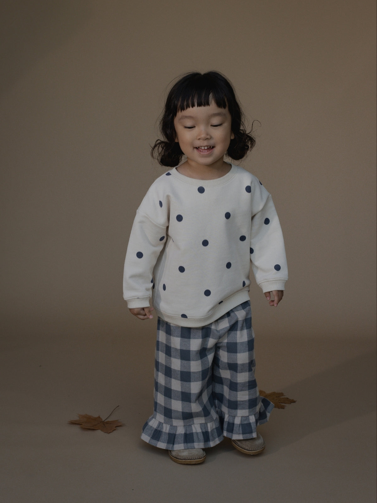 A toddler girl stands against a plain warm beige studio background wearing a cream long-sleeve top with dark blue polka dots and blue-and-white gingham wide-leg pants.