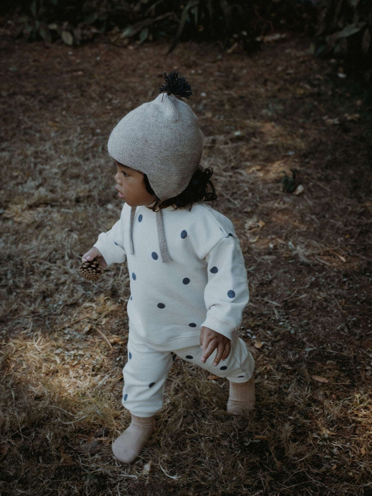 A toddler wearing a cream-colored polka dot jumpsuit, matching knit socks, and a light gray knit pom-pom hat walks through dry grass outdoors.