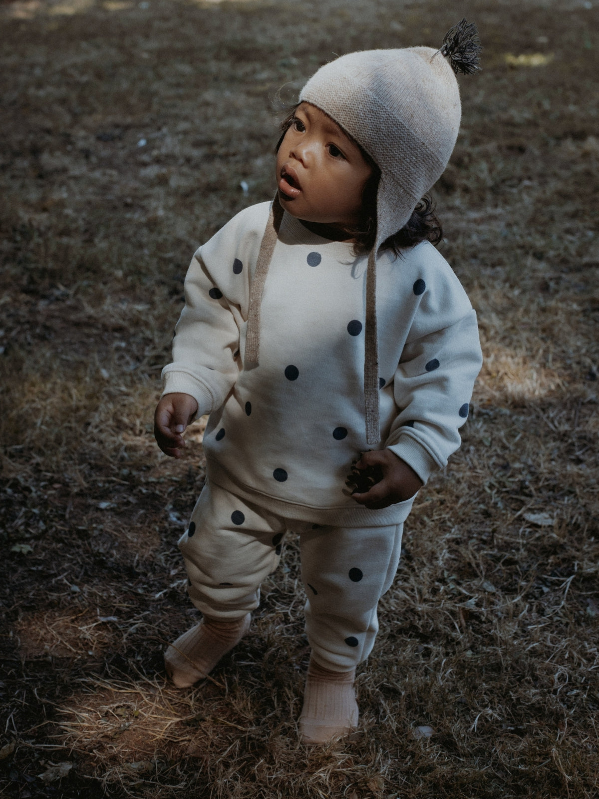 A toddler standing outdoors on dry grass wearing a cream-colored knit outfit with black polka dots, including a matching beanie with a pom-pom.