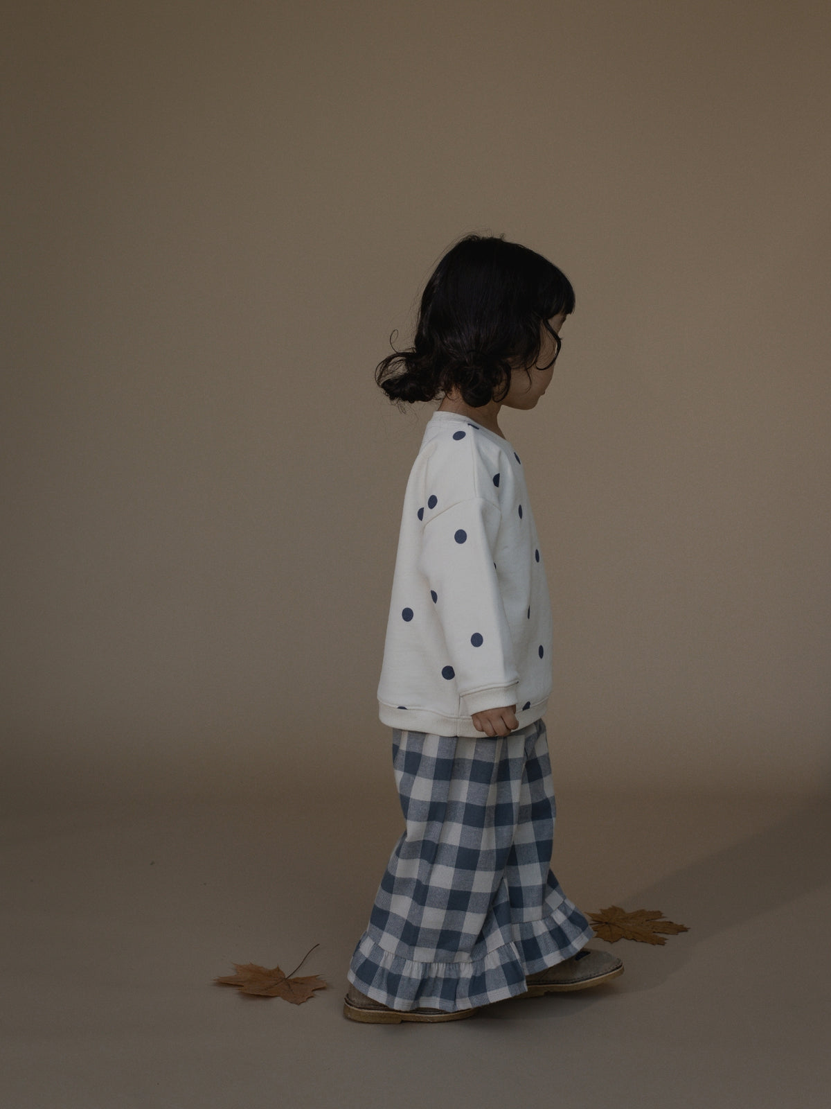 A child standing in profile wearing a white long-sleeve top with dark blue polka dots and blue-and-white gingham pants, photographed against a plain warm beige studio background.