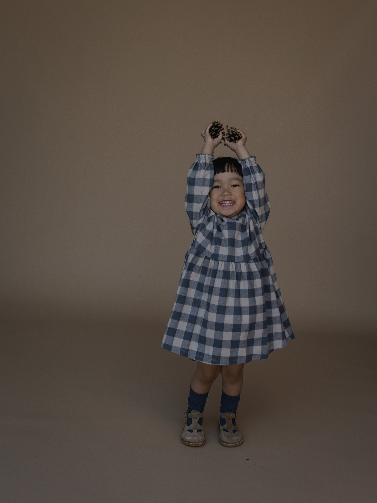 A young child wearing a blue and white gingham dress stands against a plain warm brown studio backdrop, holding binoculars up to their face.