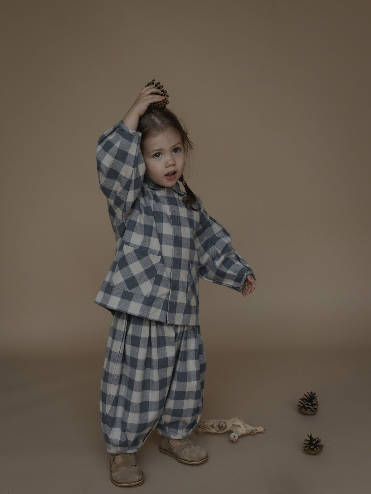 A toddler stands in a studio wearing a matching long-sleeve plaid pyjama set in muted blue-gray and off-white. The child is posed against a solid warm taupe backdrop with small pinecones and a plush toy on the floor.