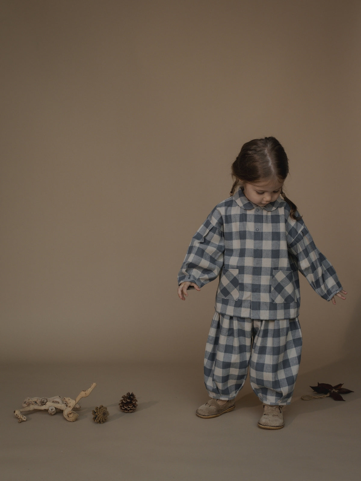 A young child wearing a blue and white gingham check pajama set with a matching long-sleeve top and pants, standing in a studio setting against a solid beige background.