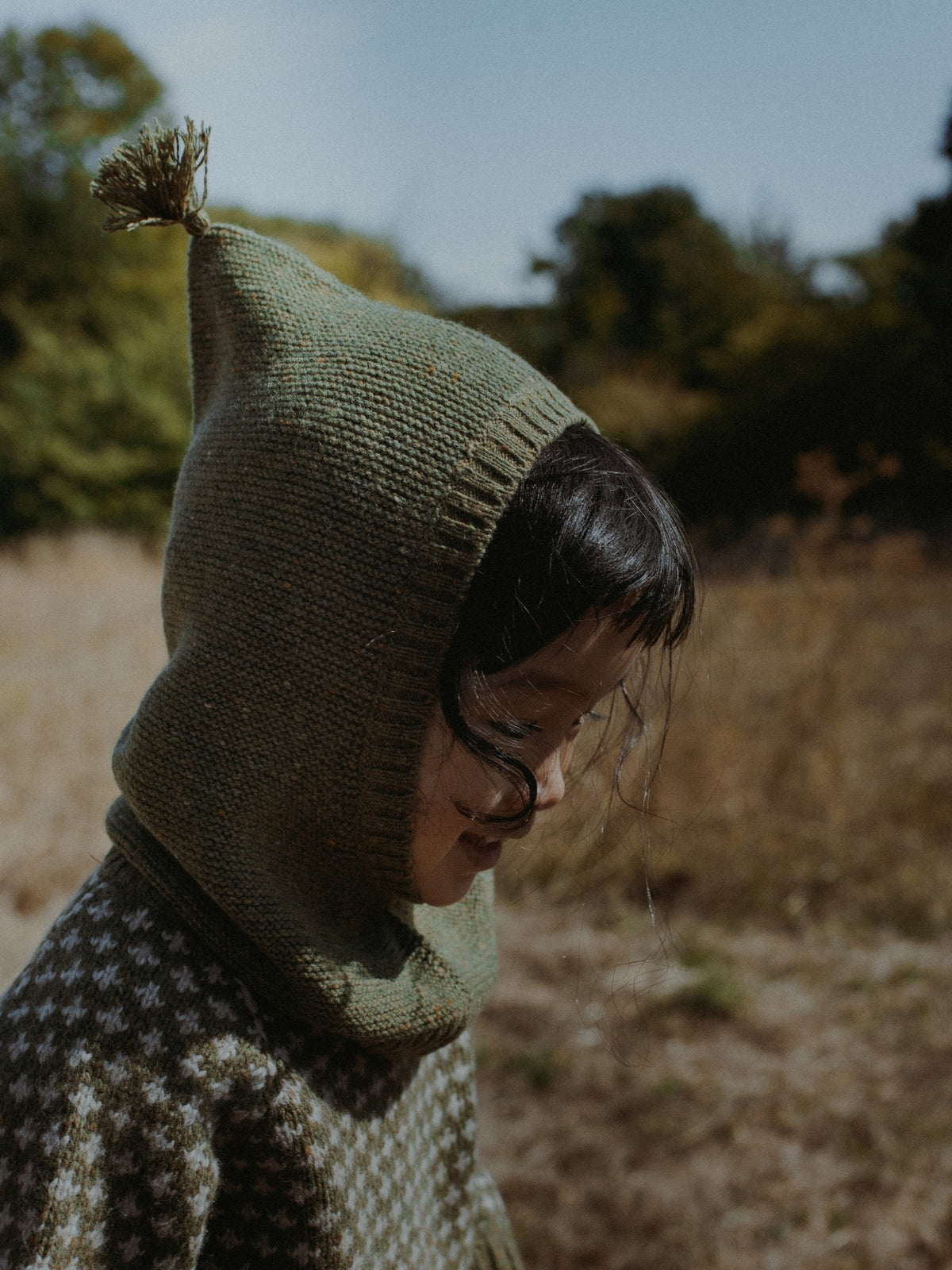 A child wearing a textured olive green knit balaclava with a small top tassel, paired with a patterned knit sweater, photographed outdoors in a dry grassy field.