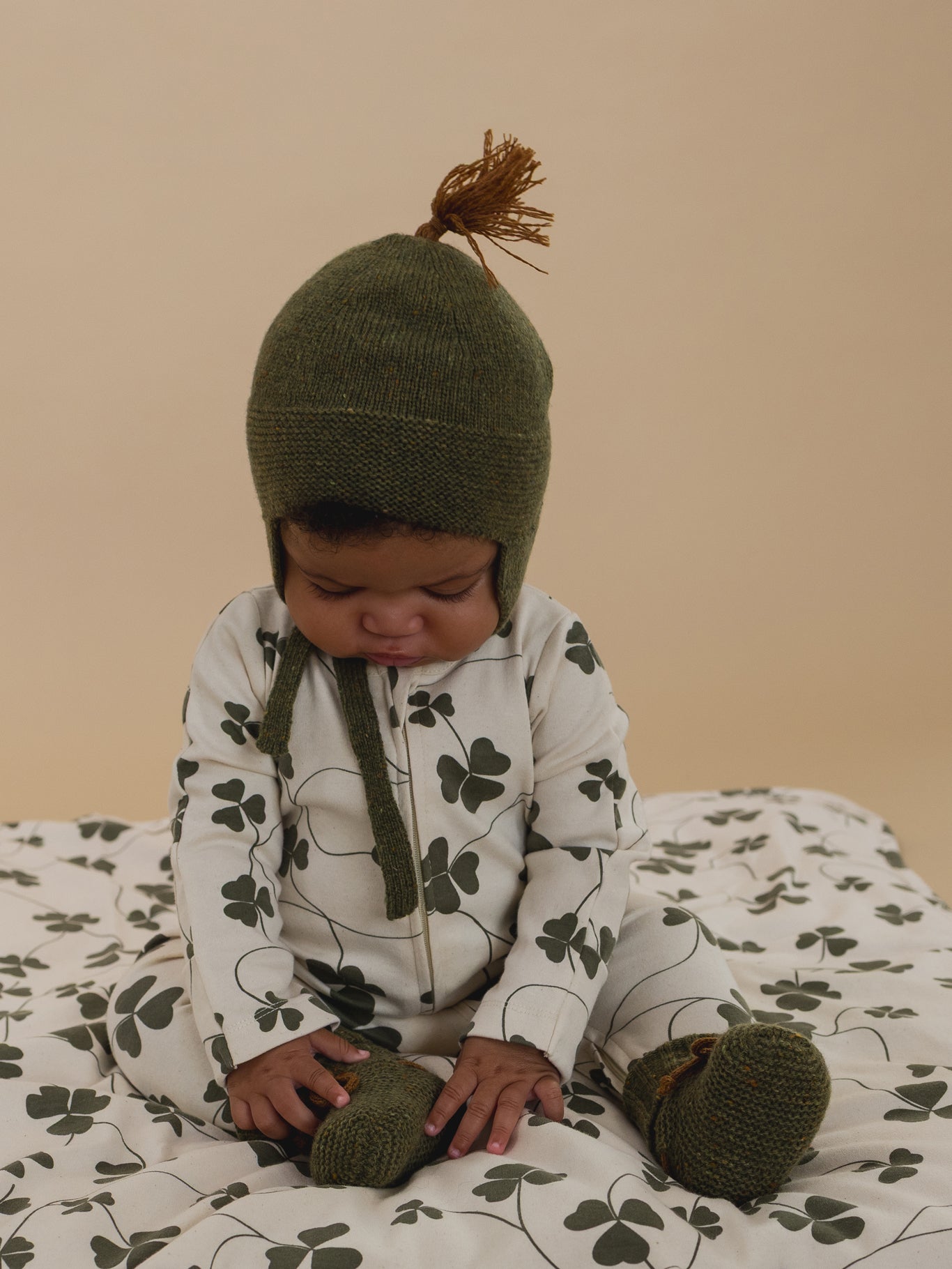 baby in forest wool tassel hat with matching booties, and clover sleepsuit holding feet