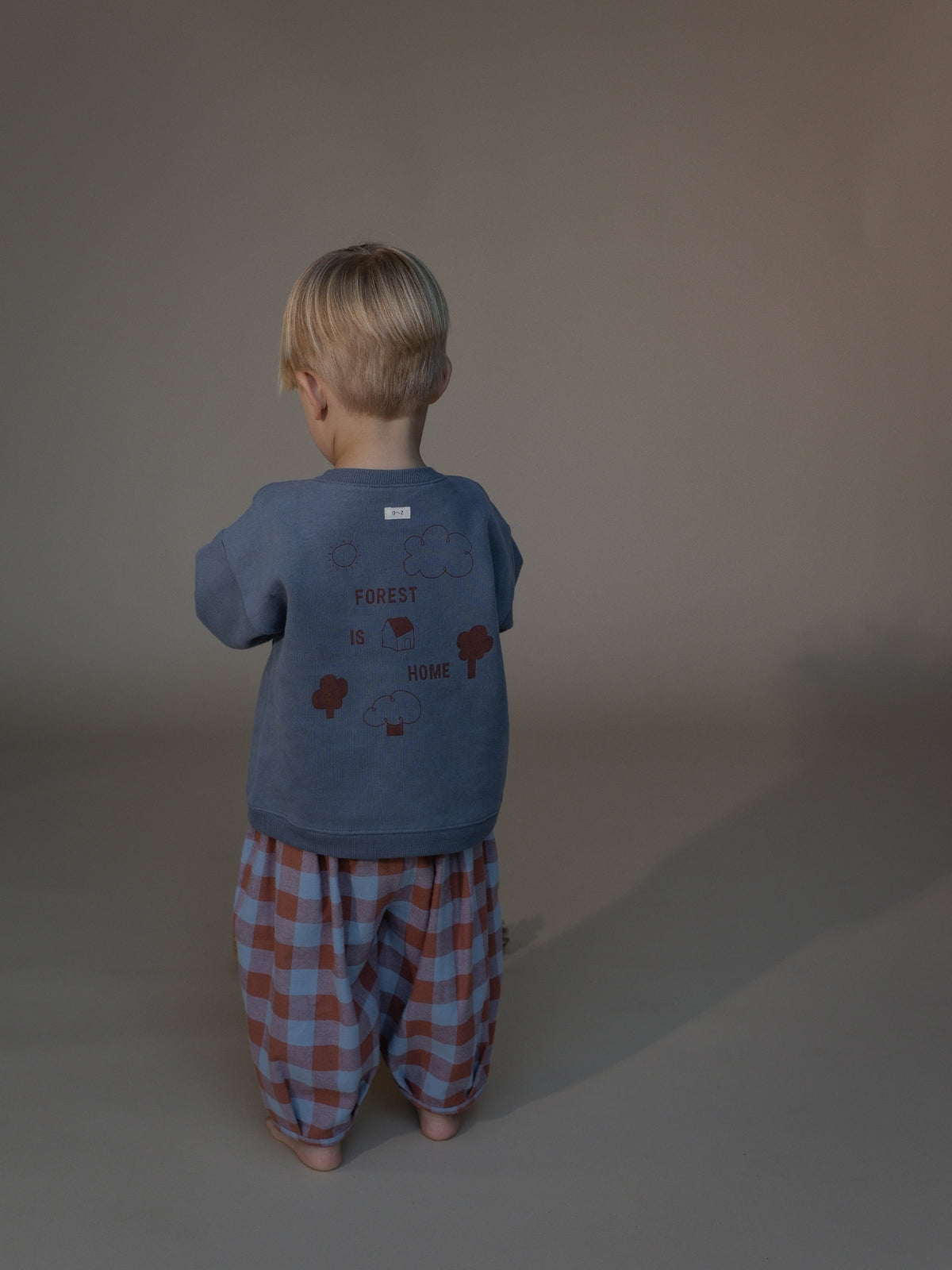 A toddler stands with their back to the camera wearing a slate blue t-shirt with a graphic print and red-and-blue checkered pants, set against a plain warm gray studio background.