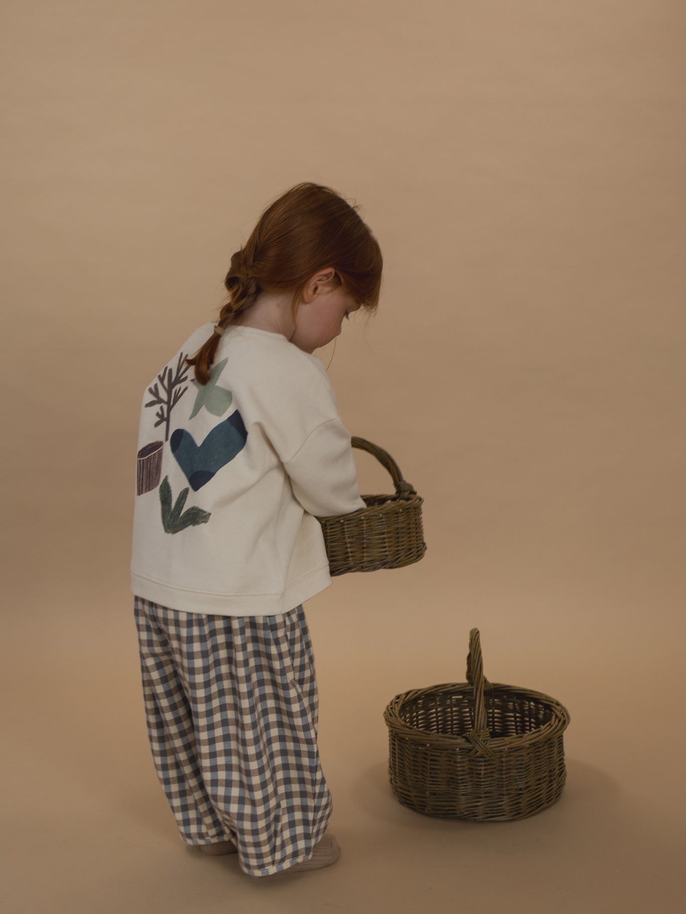 toddler in cotton sweatshirt with nature-themed back graphic and gingham pants, holding rattan basket