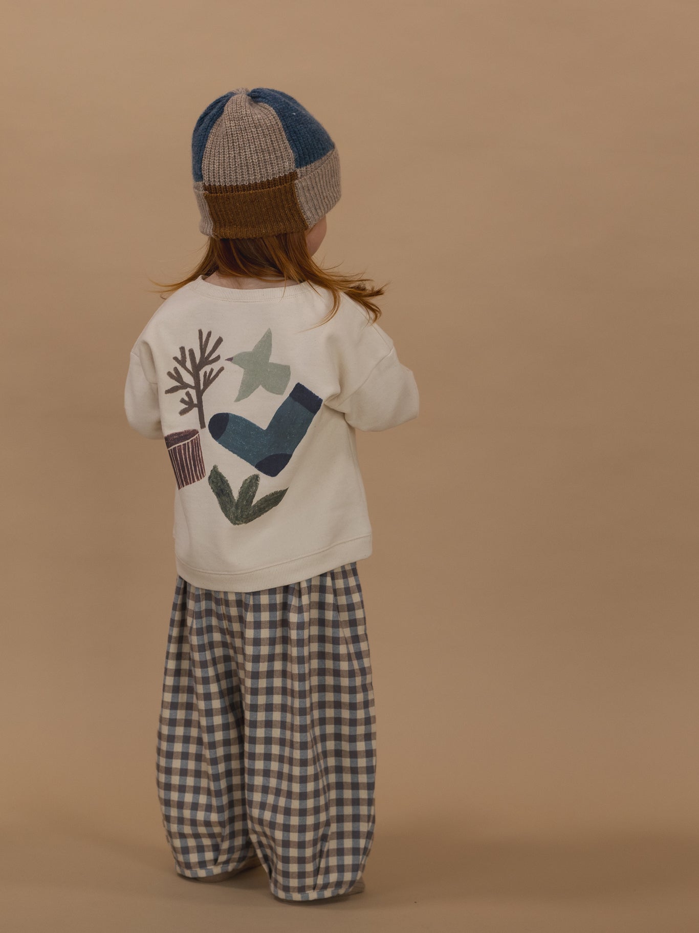 toddler in sweatshirt with nature-themed graphics on the back, gingham balloon pants and patchwork beanie