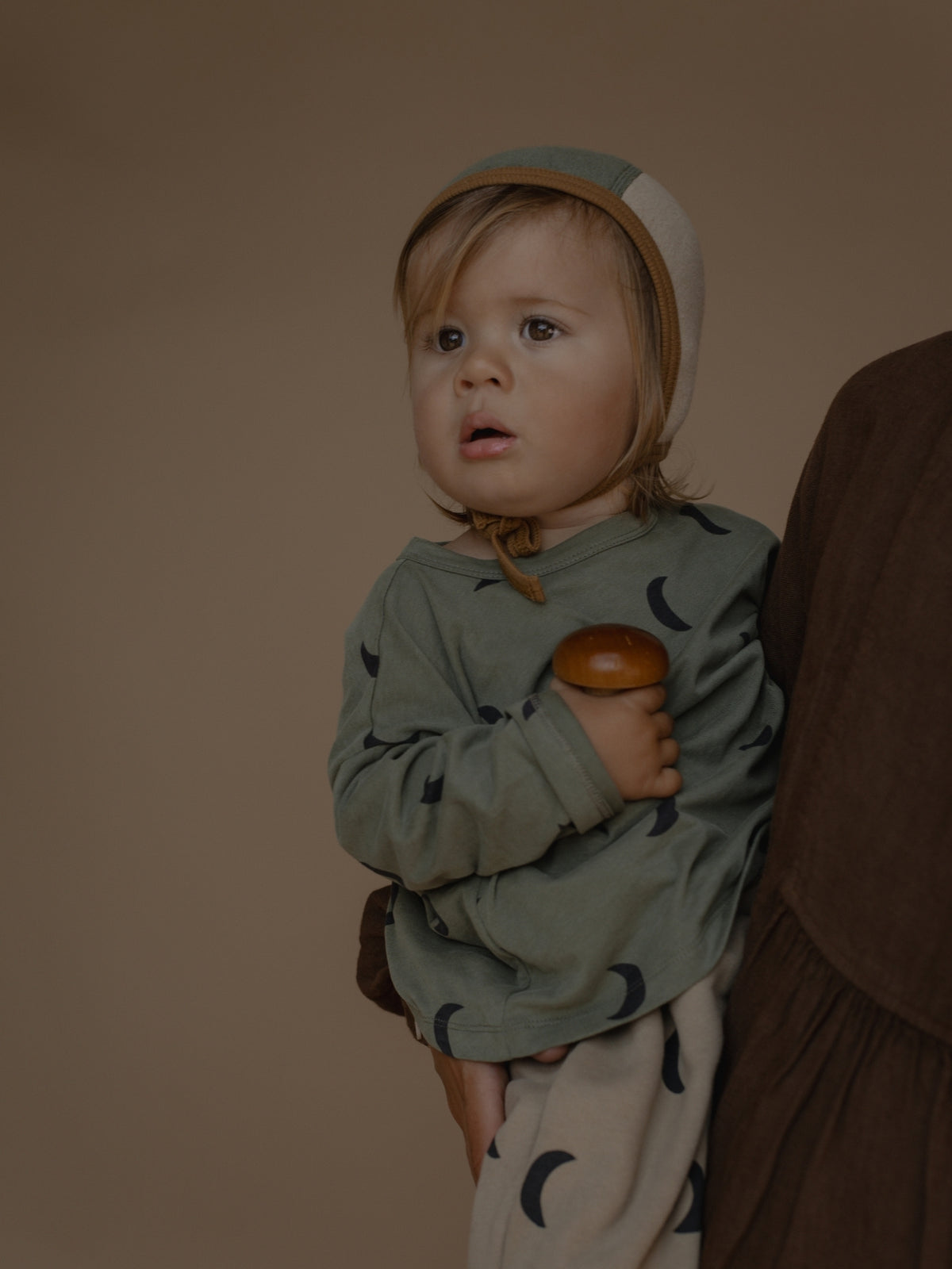 A baby wearing a sage green long-sleeve top with a dark crescent moon pattern and a matching bonnet, held by an adult wearing a brown dress against a solid brown background.