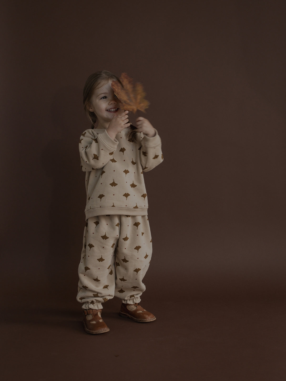 A young child wearing a cream-colored pajama set with a brown pattern, holding a dried brown leaf in front of their face, set against a solid dark brown studio background.