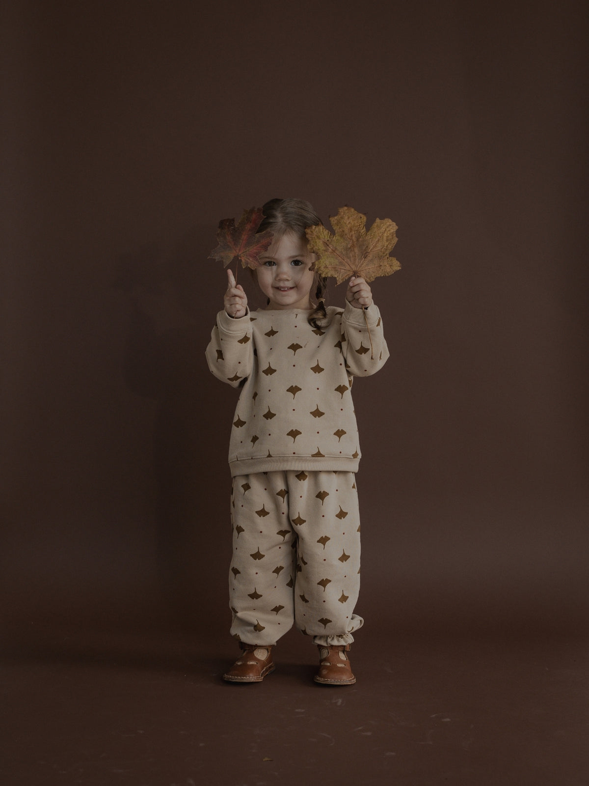 A young child stands against a dark brown studio backdrop wearing a cream-colored two-piece pajama set with a brown leaf pattern. The child holds two large autumn leaves in front of their face and wears brown shoes.