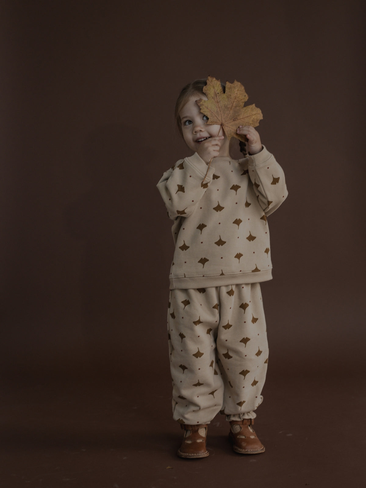 A toddler stands against a dark brown studio background wearing a cream-colored pajama set with a brown autumn leaf pattern. The child is holding a large dry leaf in front of their face.