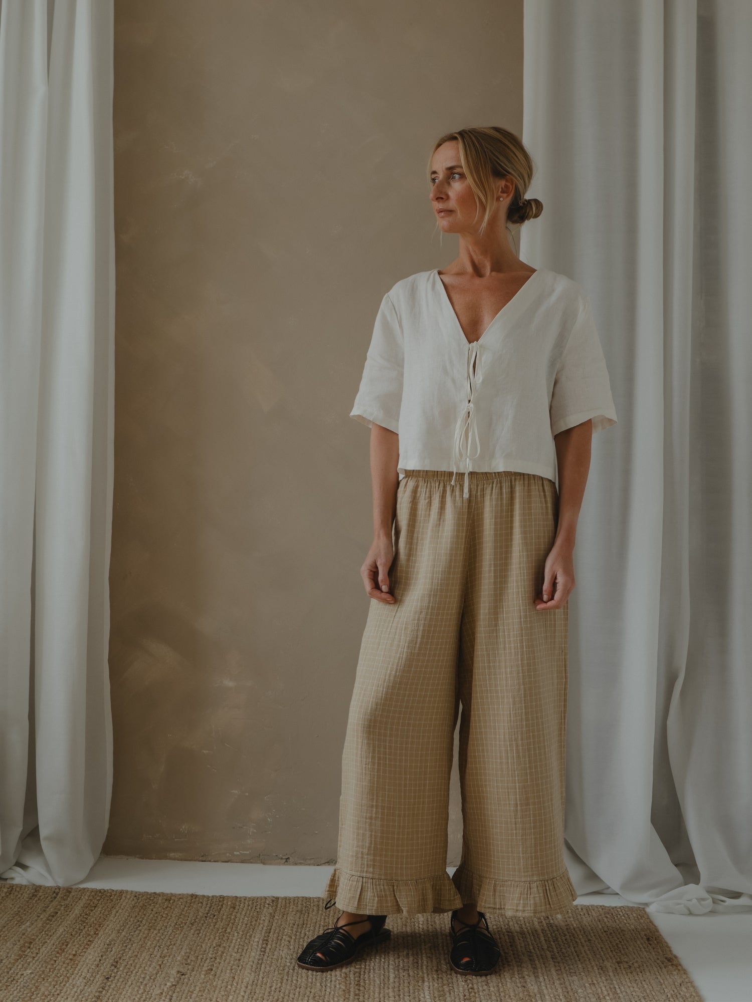 Woman wearing a white blouse and beige pants standing in front of a beige wall with white curtains.