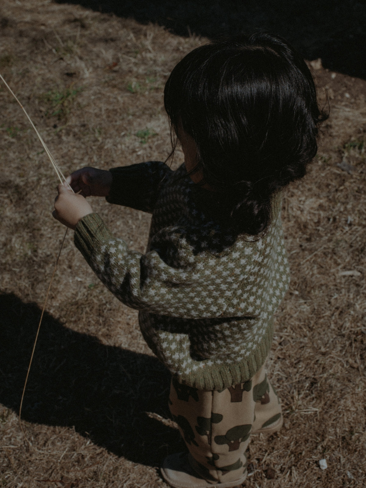 A child standing outdoors on dry grass, seen from behind, wearing a dark knit sweater with a light geometric pattern and tan pants with dark animal motifs.