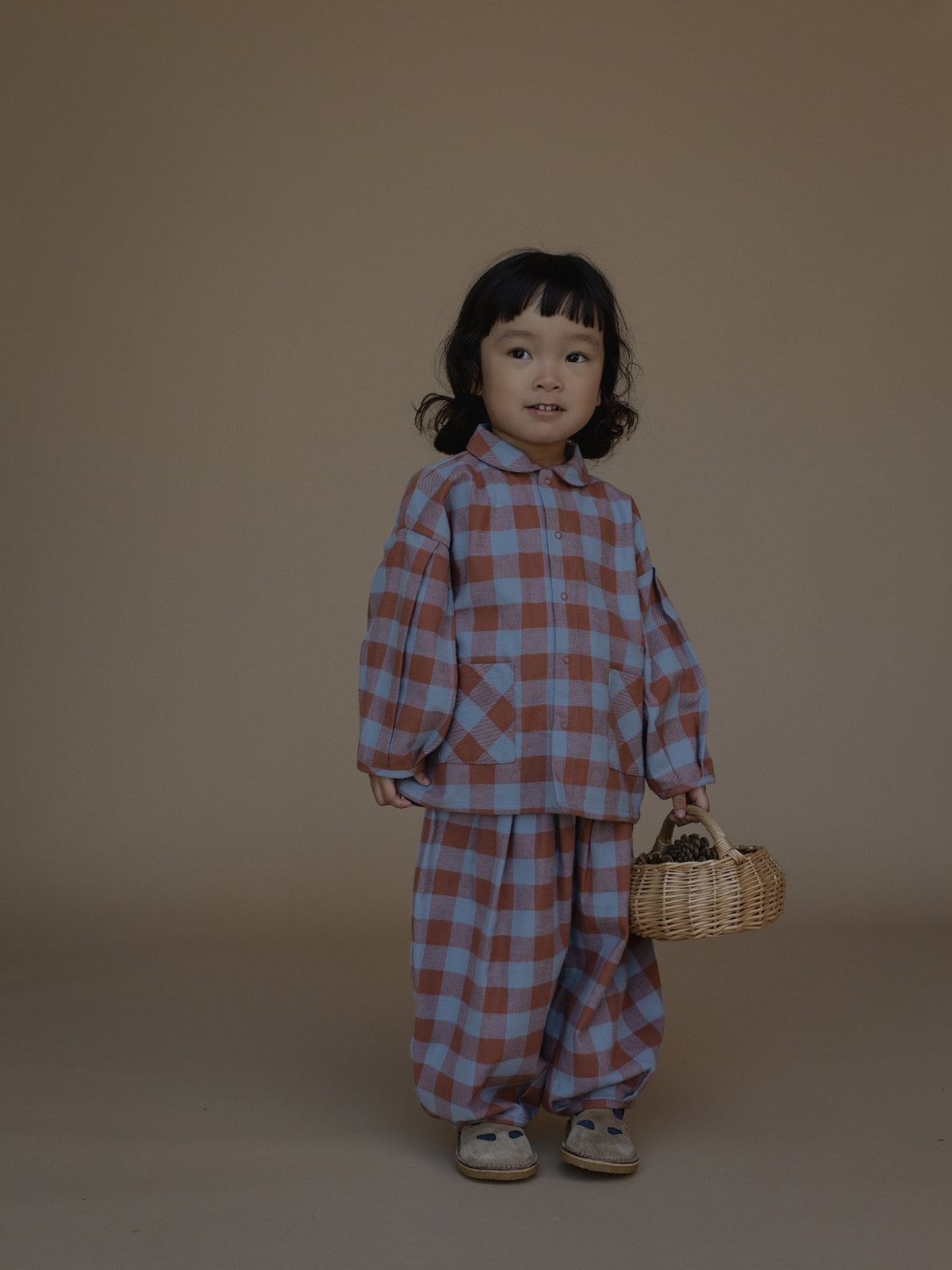 A toddler stands against a plain warm beige studio background wearing a matching plaid outfit in muted dusty blue and terracotta red, holding a small woven basket.